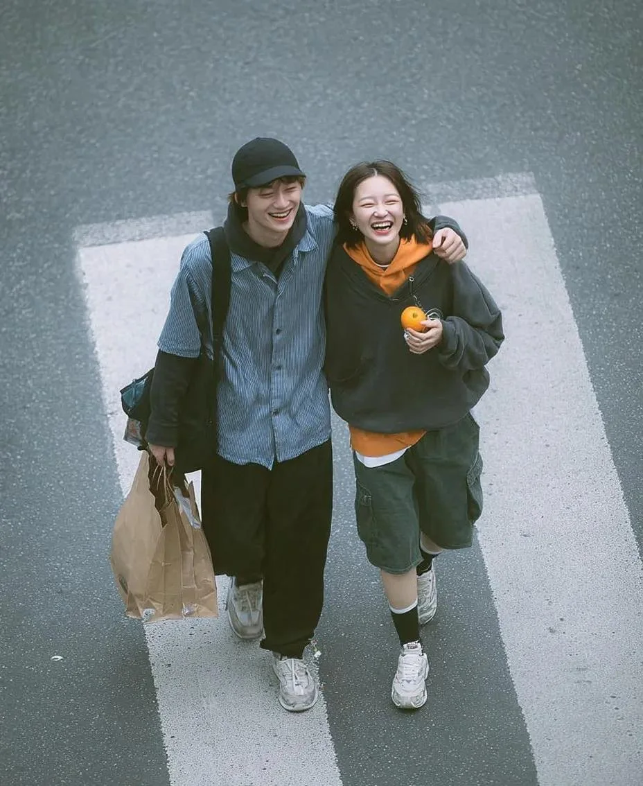 A high-angle cinematic street photo of a laughing young couple walking together on a zebra crossing, with the boy carrying a brown paper shopping bag and the girl holding a small orange fruit.