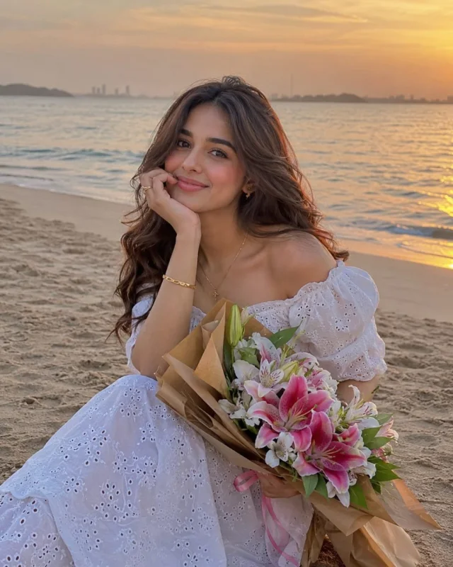 A warm sunset beach lifestyle portrait of a smiling young woman in an off-shoulder white eyelet summer dress, resting her chin on her hand while holding a wrapped bouquet of pink and white lilies on the sand with ocean waves and golden dusk sky behind her