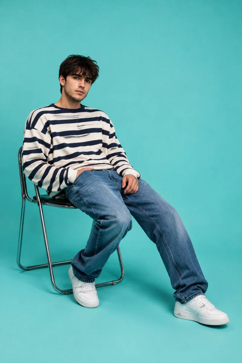 A 2:3 menswear editorial of a seated young man in a striped sweater and blue jeans against a bright aqua studio backdrop.