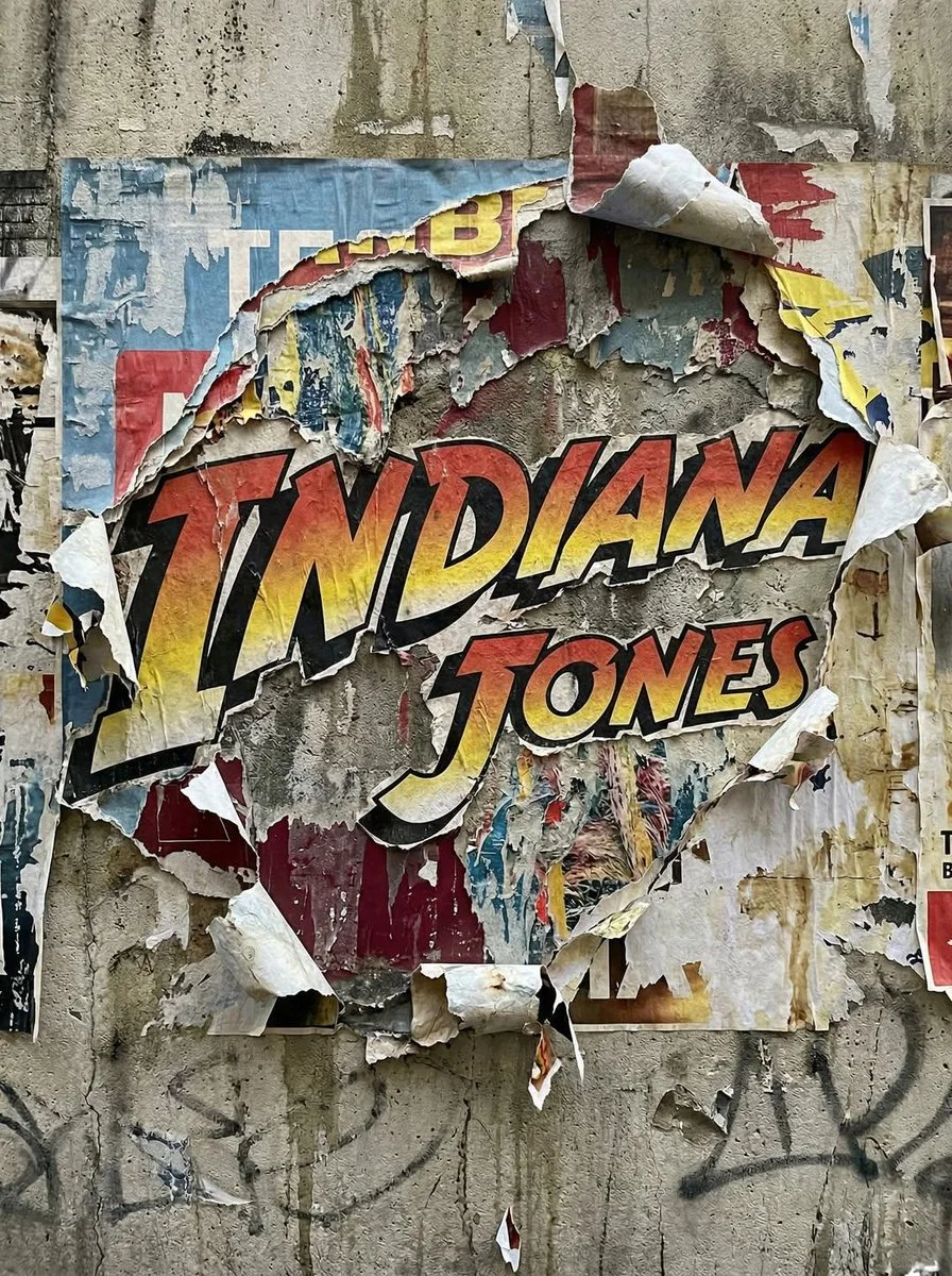 An Indiana Jones logo exposed beneath layered torn street posters, with fibrous edges, peeling paper, concrete wall texture, and urban wear.