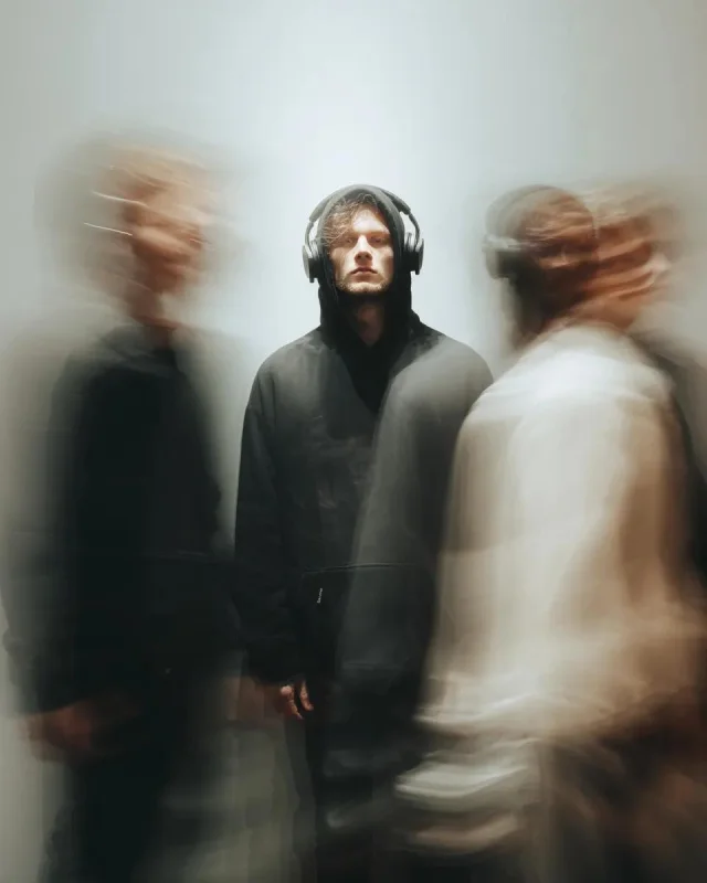 A solitary young man in a black hoodie and headphones stands sharply still while blurred pedestrians rush past around him.