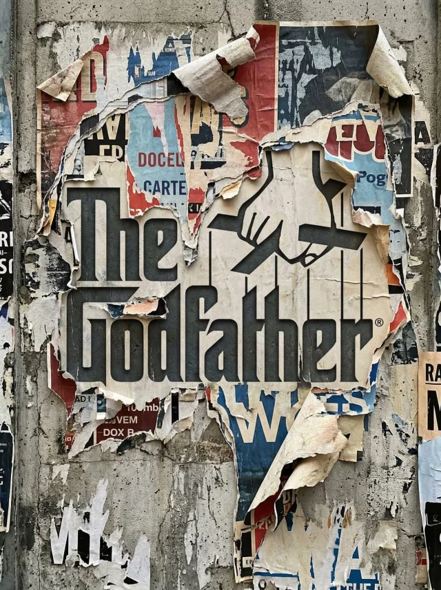 A realistic urban concrete wall with layers of ripped posters revealing The Godfather logo silhouette through aged paper, curled torn edges, stains, and glue residue.