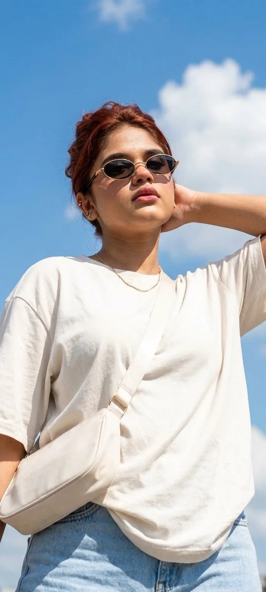 A second lower-angle summer-sky portrait of the same woman with gold sunglasses and white tee.