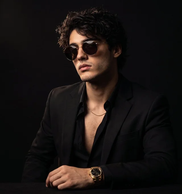 A low-key editorial fashion portrait of a young man in a black suit and open black shirt, rounded gold sunglasses, gold chain and watch, seated against a near-black studio background with chiaroscuro lighting.