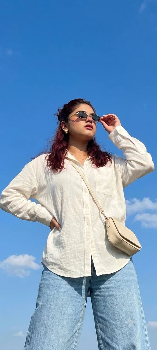 A lower-angle portrait of a woman against a clear summer sky, oval gold-frame sunglasses, white oversized tee, hand behind head.