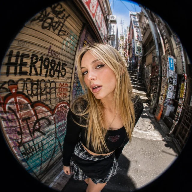 A square ultra-wide-angle fisheye portrait of a blonde model in a narrow steep Tokyo graffiti alley, with heavy barrel distortion, black circular vignette, sharp eyes, metal shutters covered in graffiti, and bright midday light.