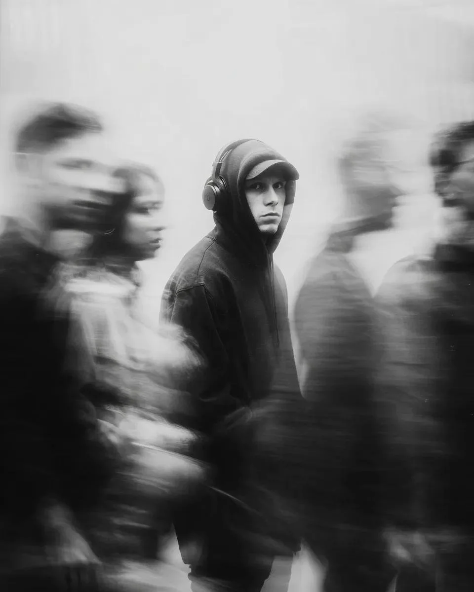 Cinematic psychological portrait variation with a hooded man frozen in focus amid ghostly long-exposure crowd motion.