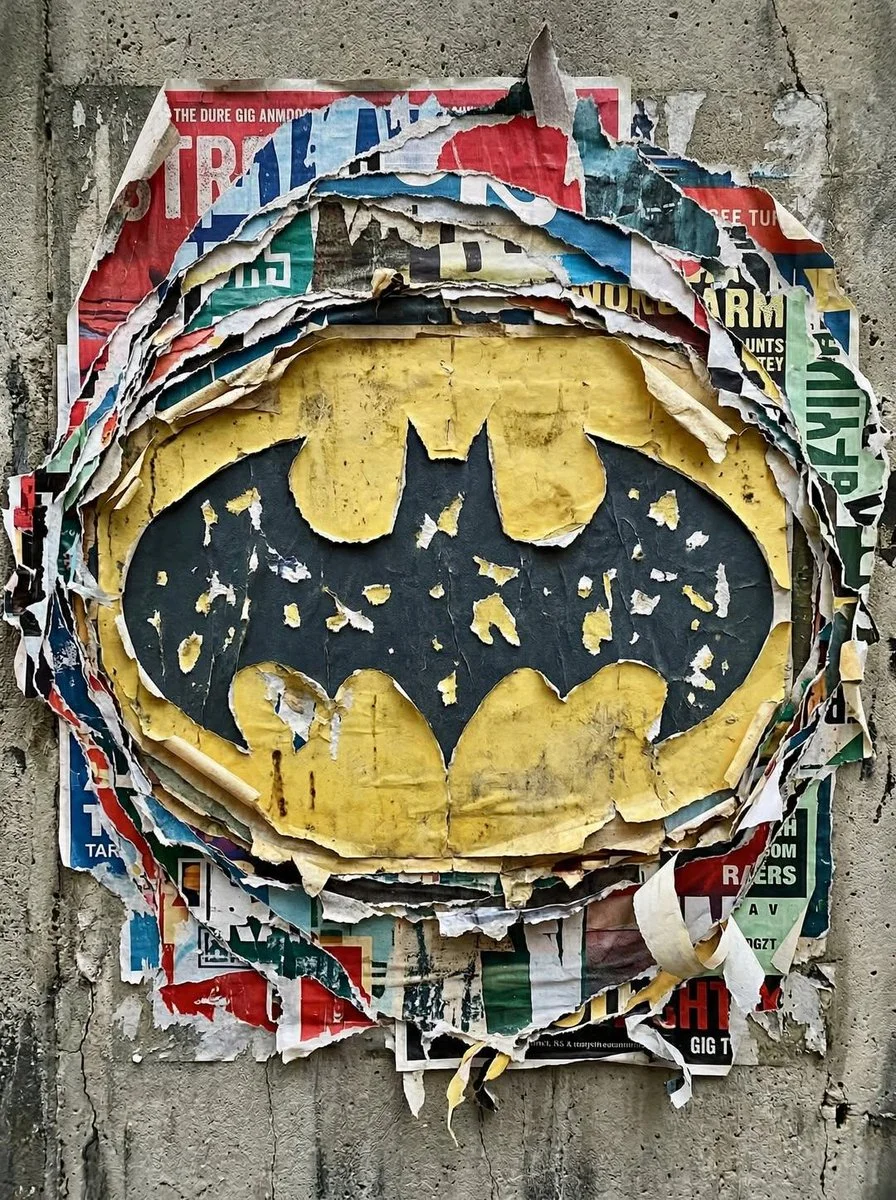 A Batman logo revealed through layered ripped posters on a rough wall, surrounded by curled paper fragments, dirt, weathering, and graffiti-like street texture.